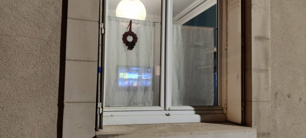 La fenêtre du salon vue de la rue, l'écran de la TV visible à travers le voilage